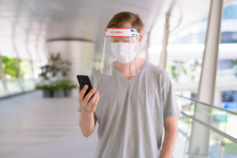 Young man using phone with mask and face shield for protection from corona virus outbreak in the city outdoors Young man using phone with mask and face shield for protection from corona virus outbreak in the city outdoors